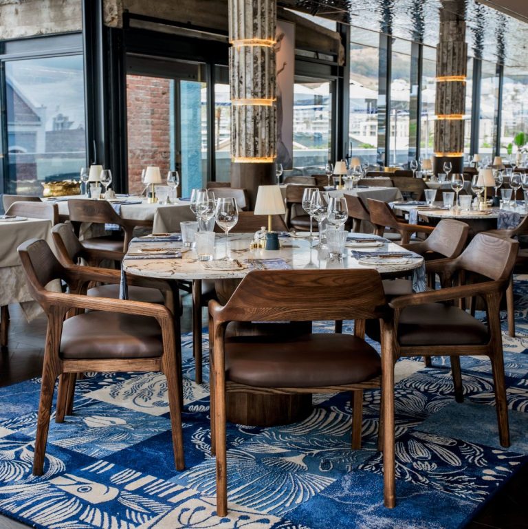 An elegant dining room with a view of the harbor, featuring a bold blue patterned rug and stylish wooden chairs at Marble's rooftop bar in Cape Town