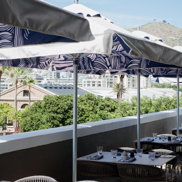 A scenic outdoor terrace with set tables and stylish umbrellas at Marble's rooftop bar at the V&A Waterfront, offering a panoramic view of the city